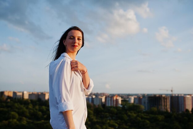 Retrato de una mujer destacada con camisa blanca masculina posando en la parte superior del edificio con un paisaje encantador en el fondo