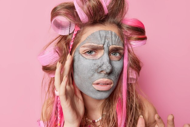 Retrato de mujer disgustada toca la cara aplica máscara de arcilla de belleza frunce el ceño la cara usa rodillos para el cabello se siente infeliz cuida la tez aislada sobre fondo rosa Procedimientos antienvejecimiento