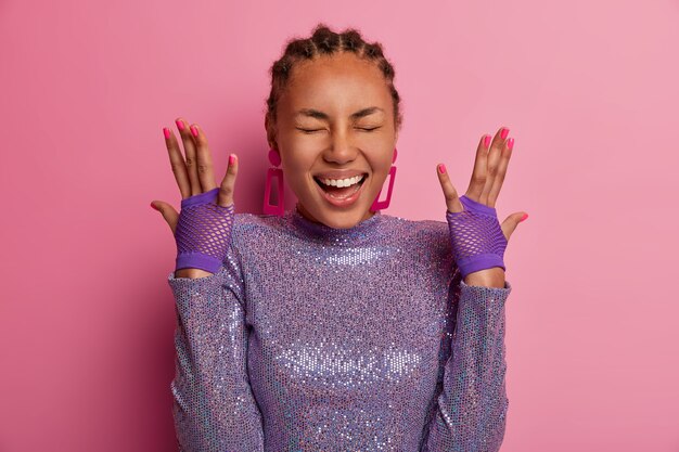 El retrato de una mujer divertida muy feliz tiene la piel oscura, levanta las manos, muestra guantes deportivos, usa un jersey púrpura brillante, se ríe positivamente, escucha algo hilarante, modela sobre una pared rosa pastel