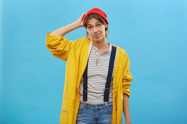 Retrato de mujer dudosa vestida casualmente rascándose la cabeza con la mano sin saber qué hacer aislado sobre la pared azul. Bonita mujer en anorak suelto amarillo que no está seguro de tener dudas