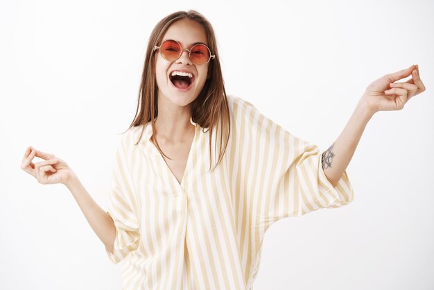 Retrato de mujer elegante despreocupada feliz y divertida alegre en gafas de sol rojas de moda y blusa de rayas amarillas bailando cantando canciones en voz alta con una amplia sonrisa y las manos extendidas a un lado