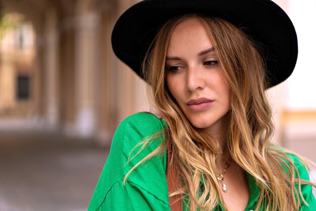 Retrato de mujer elegante elegante con cabello rizado rubio y maquillaje natural de cerca, con sombrero negro posando en la calle