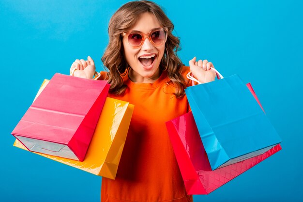 Retrato de mujer elegante sonriente atractiva excitada adicta a las compras en vestido de moda naranja con bolsas de compras sobre fondo azul de estudio aislado