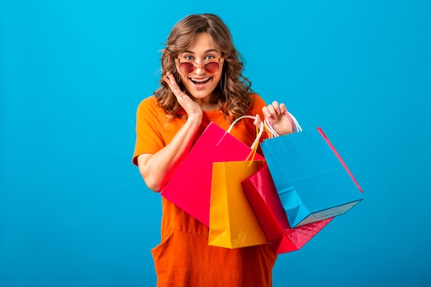 Retrato de mujer elegante sonriente atractiva excitada adicta a las compras en vestido de moda naranja con bolsas de compras sobre fondo azul de estudio aislado
