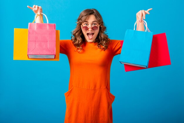 Retrato de mujer elegante sonriente atractiva excitada adicta a las compras en vestido de moda naranja con bolsas de compras sobre fondo azul de estudio aislado