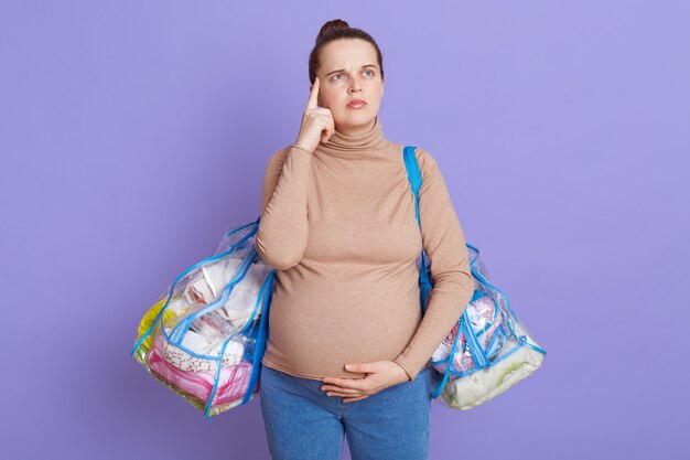 Retrato de mujer embarazada pensativa, tratando de recordar que lleva todo al hospital, mantiene el dedo en la sien, mirando hacia arriba con expresión pensativa, aislado sobre una pared lila