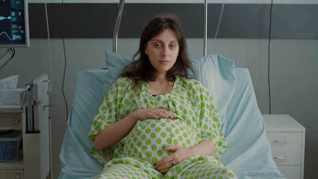 Retrato de mujer embarazada tendido en la cama de la sala de hospital con equipo médico preparándose para el parto en la maternidad. Paciente caucásico sosteniendo la mano sobre el vientre mientras está sentado esperando bebé