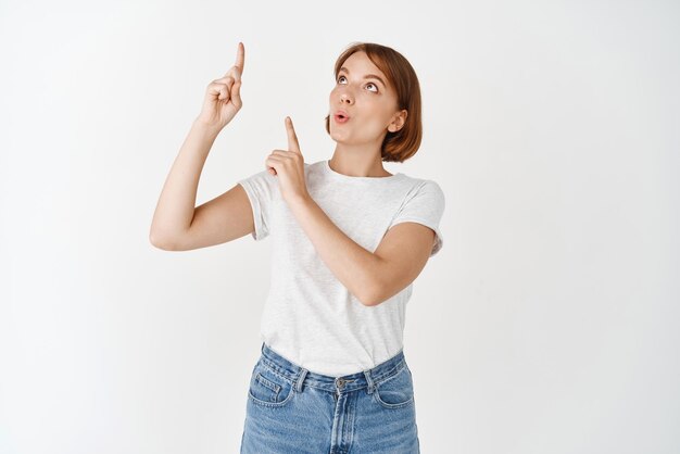 Retrato de una mujer emocionada con una camiseta que señala y mira hacia arriba divertida al ver una pancarta interesante que demuestra algo en el espacio de una copia en blanco