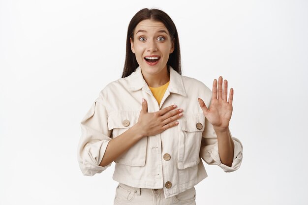 Retrato de mujer emocionada se ve sorprendida, levanta la mano y pone el brazo en el pecho, dice la verdad, la palabra honesta, saluda, se presenta, de pie con ropa casual en blanco.