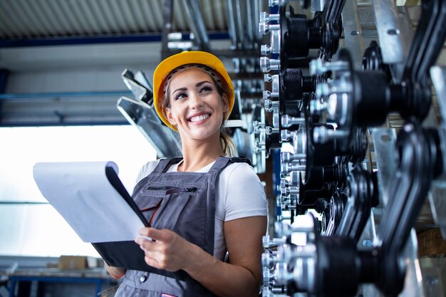 Retrato de mujer empleada industrial en uniforme de trabajo y casco comprobando la producción en la fábrica.