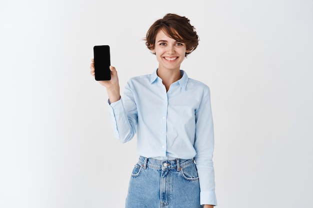 Retrato de mujer empresaria sonriendo y mostrando la pantalla vacía del teléfono inteligente, de pie con camisa de cuello azul en la pared blanca