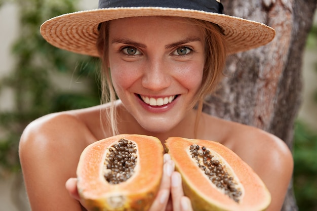 Retrato de mujer encantadora alegre posa desnuda, usa sombrero de verano, sostiene papaya exótica orgánica