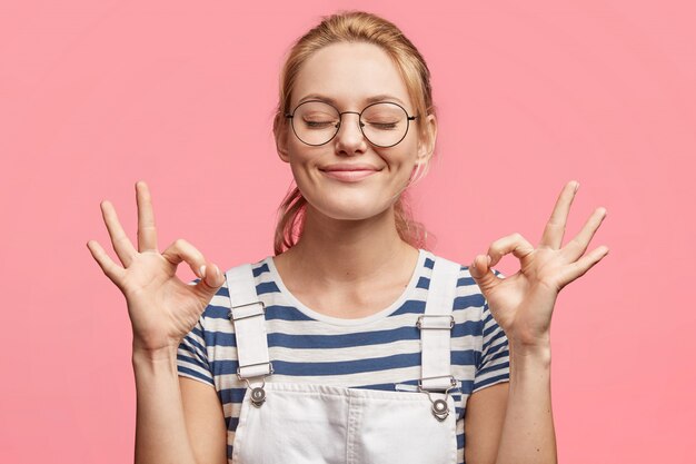 Retrato de mujer encantadora atractiva rubia satisfecha indica signo de ok, mantiene los ojos cerrados, está de buen humor, feliz de tener fines de semana, aislado sobre rosa