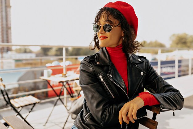 Retrato de mujer encantadora elegante de boina roja con labios rojos sentado en la terraza al aire libre de cerca