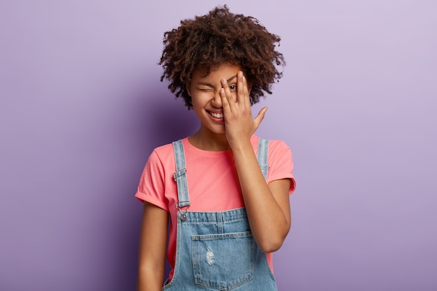 Retrato de mujer encantadora feliz cubre la cara con la palma, mira a través de los dedos, parpadea, sonríe ampliamente, intenta esconderse