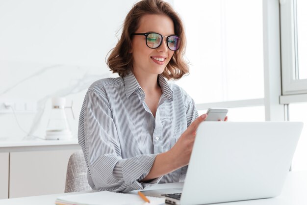 Retrato de mujer encantadora en gafas y camisa a rayas con teléfono móvil mientras se encuentra en el lugar de trabajo en la sala blanca