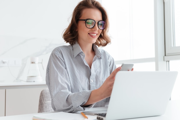 Retrato de mujer encantadora en gafas y camisa a rayas con teléfono móvil mientras se encuentra en el lugar de trabajo en la sala blanca