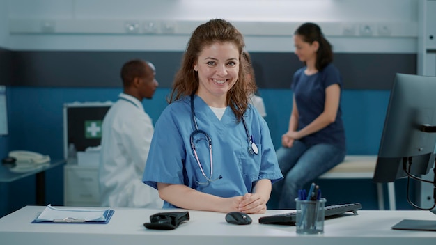 Retrato de mujer enfermera sonriendo y usando uniforme en la oficina, sentada en el escritorio. Asistente médico con estetoscopio mirando la cámara y preparándose para ayudar al médico en la cita.