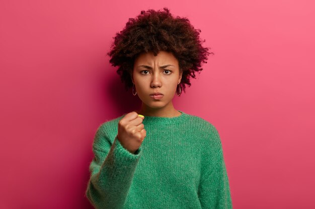 El retrato de una mujer enojada de pelo corto muestra el puño, tiene una expresión irritada, promete venganza, usa un suéter verde, posa contra la pared rosa, domina y amenaza, está insatisfecho