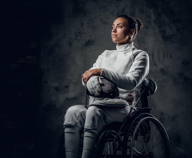 Foto gratuita retrato de mujer esgrimista en silla de ruedas con máscara de seguridad y estoque sobre fondo gris.