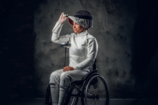 Foto gratuita retrato de mujer esgrimista en silla de ruedas con máscara de seguridad y estoque sobre fondo gris.