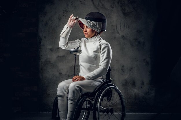 Retrato de mujer esgrimista en silla de ruedas con máscara de seguridad y estoque sobre fondo gris.