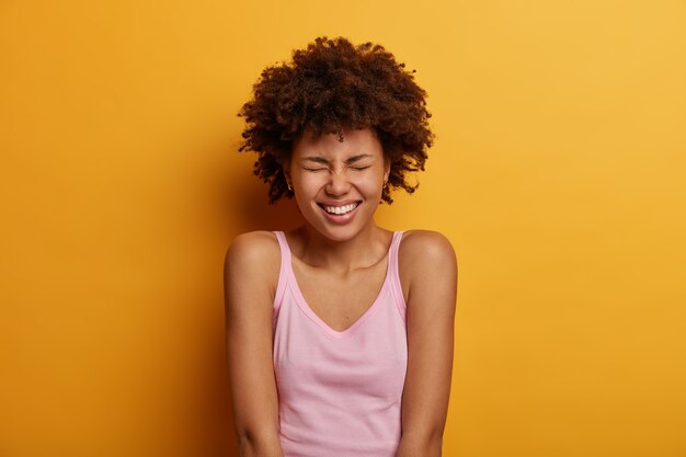 El retrato de una mujer étnica positiva entrecierra la cara, sonríe felizmente, muestra los dientes blancos, está de buen humor, disfruta del día libre, escucha los buenos chistes de un amigo, usa un chaleco informal, modelos contra la pared amarilla