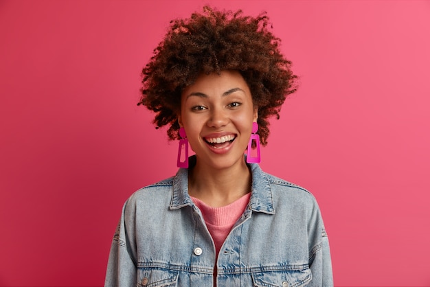 Foto gratuita el retrato de una mujer étnica satisfecha se ve con una sonrisa con dientes, tiene un estado de ánimo feliz, se ríe de una broma hilarante, se viste con un atuendo de moda, se siente despreocupada, aislada en la pared rosa