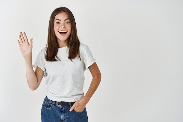 Retrato de mujer europea alegre en camiseta, agitando la mano en gesto de saludo y sonriendo ampliamente, pared blanca.