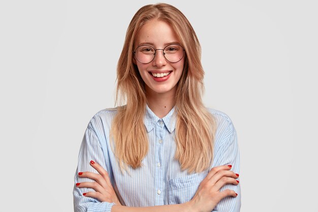 Retrato de mujer europea alegre con expresión satisfecha, vestida con camisa y gafas, se regocija al recibir una buena nota, aislada sobre una pared blanca