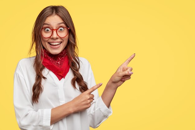 El retrato de una mujer europea alegre tiene dos trenzas, sonríe ampliamente, usa un elegante pañuelo y una camisa blanca, señala en la esquina superior derecha, modelos contra la pared amarilla, sugiere ir a este lugar