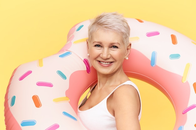 Retrato de mujer europea atractiva confiada con corte de pelo elegante de duendecillo posando sobre fondo amarillo, llevando un círculo de natación inflable rosa, pasando un día soleado junto al mar o en la piscina