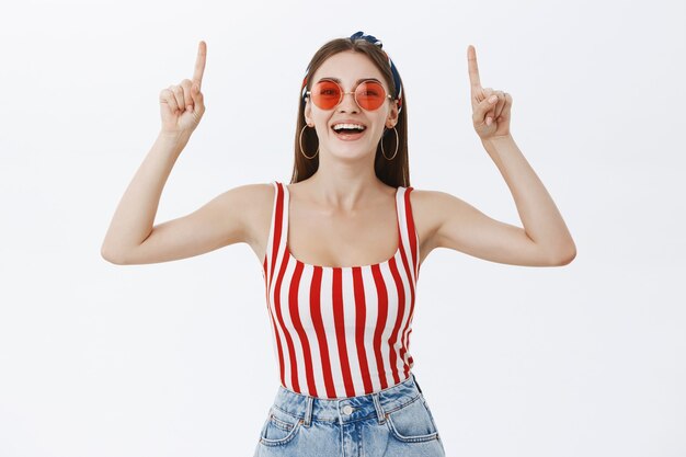 Retrato de mujer europea elegante con pelo largo en la parte superior de rayas de moda y gafas de sol levantando la mano apuntando hacia arriba y sonriendo divertido y encantado