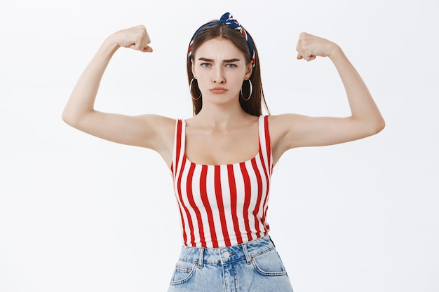 Retrato de mujer europea fuerte y segura de buen aspecto con estilo en la parte superior a rayas y diadema frunciendo los labios y frunciendo el ceño haciendo una cara seria que muestra los músculos y bíceps