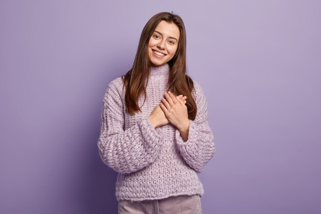 Retrato de mujer europea joven feliz mantiene las manos en el pecho, muestra el gesto del corazón, expresa gratitud, estar agradecido, modelos contra la pared púrpura Lenguaje corporal. Monocromo. Gente y devoción