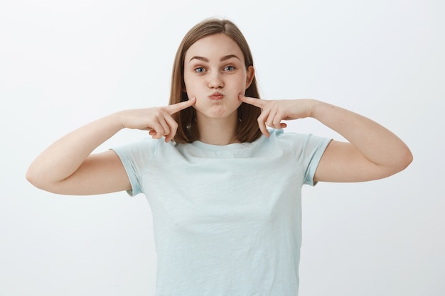 Retrato de mujer europea linda despreocupada en camiseta azul claro casual haciendo pucheros, conteniendo la respiración y tocando las mejillas con los dedos índices divirtiéndose pasando tiempo sobre la pared gris tratando de divertirse