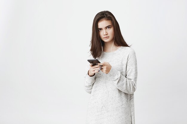 Retrato de mujer europea linda preocupada y frustrada, sosteniendo teléfono inteligente y usando auriculares