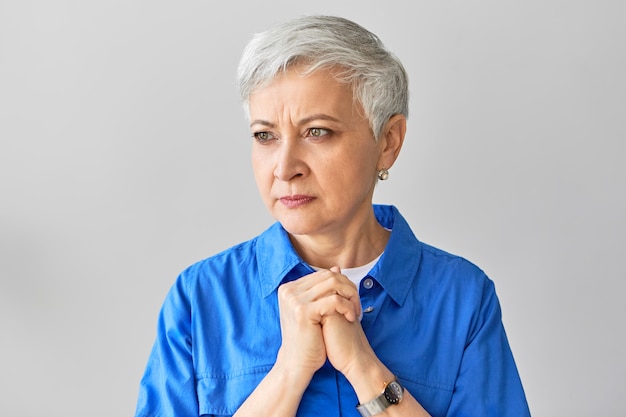 Foto gratuita retrato de mujer europea madura de mediana edad con el ceño fruncido serio con cabello gris de duendecillo que expresa nerviosismo, sosteniendo las manos juntas sobre su pecho, siendo impaciente, esperando los resultados del análisis de sangre