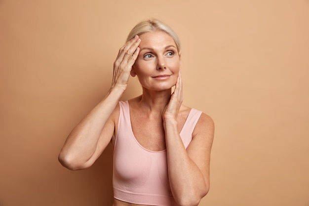 Retrato de una mujer europea mayor madura toca la cara suavemente tiene una piel perfecta y mira pensativamente lejos disfruta de su tez suave se preocupa por la apariencia satisfecha después del procedimiento anti-envejecimiento