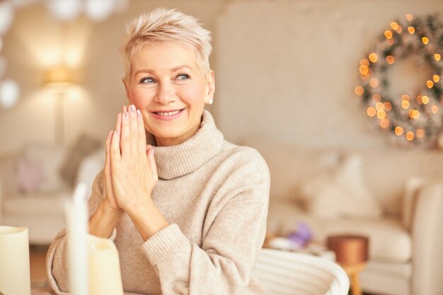 Retrato de mujer europea de mediana edad atractiva feliz con maquillaje cuidado y elegante corte de pelo sentado en la sala de estar con guirnaldas y guirnaldas hechas a mano de pino