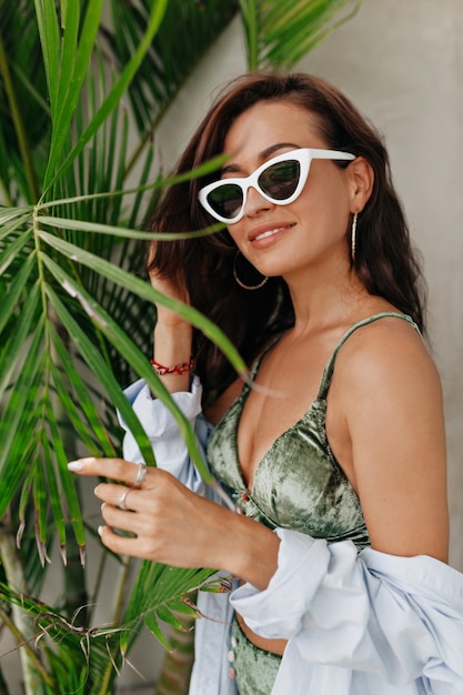 Retrato de mujer europea de moda elegante en traje de moda con pelo largo con camisa y gafas. Chica sexy posando por hojas de palma.