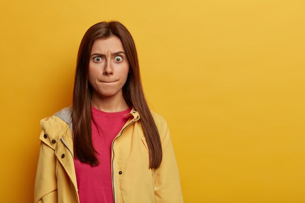 Retrato de mujer europea preocupada emocionalmente presiona los labios, intenta controlar sus emociones, levanta las cejas, viste un jersey rosa y un anorak