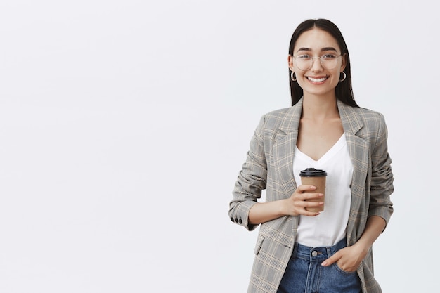 Retrato de mujer europea relajada y segura con cabello oscuro y gafas, sosteniendo la mano en el bolsillo y bebiendo té