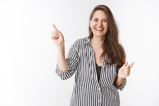 Retrato de una mujer feliz, activa y saludable, de mediana edad, con blusa a rayas, sonriendo divertida, divirtiéndose, apuntando hacia arriba y alegremente satisfecha con la calidad del producto sobre fondo blanco.