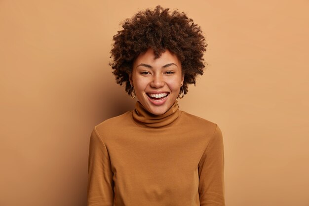 Retrato de mujer feliz y alegre de piel oscura sonríe ampliamente, tiene dientes blancos perfectos, expresa felicidad, se siente afortunada durante un día positivo, usa un jersey de poloneck, posa contra una pared marrón