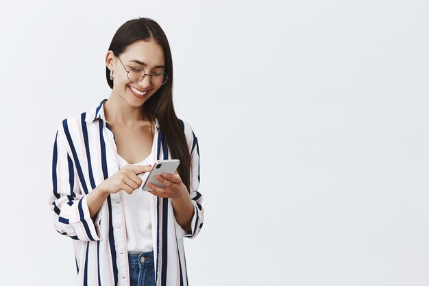 Retrato de mujer feliz atractiva y elegante con gafas y blusa a rayas, alimentación de desplazamiento mientras usa el teléfono inteligente, leyendo un artículo divertido en Internet