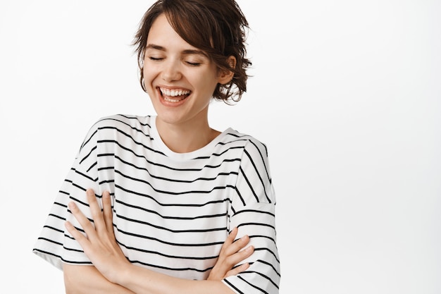 Retrato de mujer feliz atractiva riendo, brazos cruzados sobre el pecho, sonriendo con los ojos cerrados, de pie en camiseta a rayas en blanco.