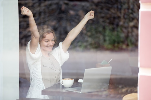 Foto gratuita retrato de mujer feliz en la cafetería celebrando el éxito con sus manos arriba