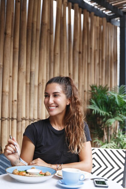 Retrato de una mujer feliz comiendo afuera desayunando y hablando con un amigo en la mesa mirando a un lado con una sonrisa alegre sentada cerca del teléfono móvil en la terraza