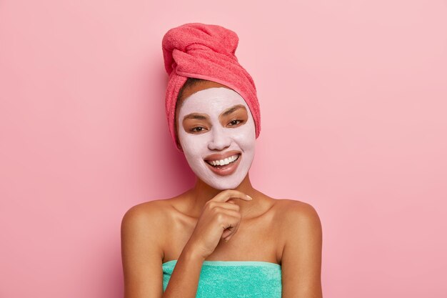 Retrato de mujer feliz y complacida con máscara facial de arcilla, inclina la cabeza y toca la barbilla, envuelta en una toalla, se preocupa por la belleza, sonríe feliz y muestra dientes blancos